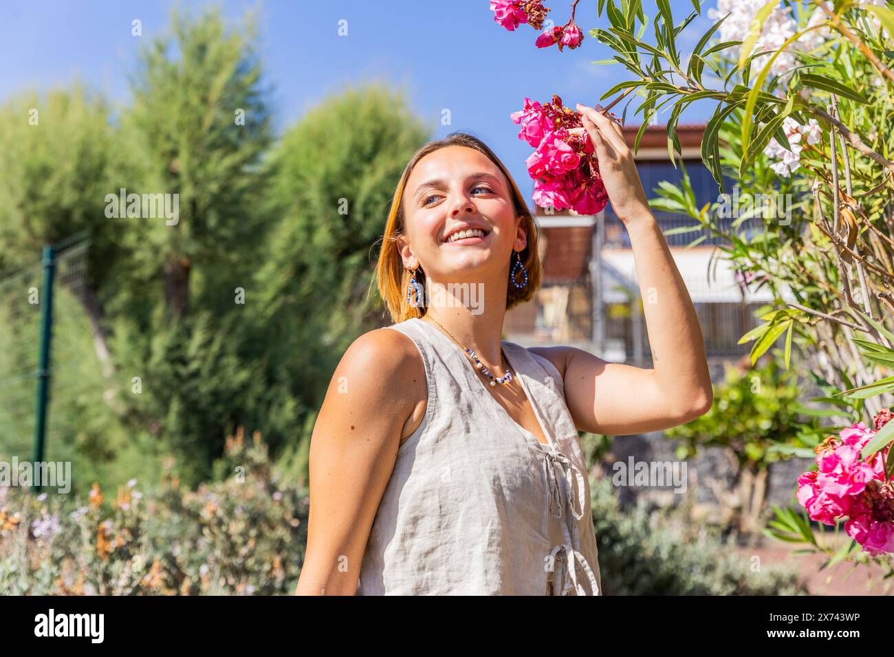 Eine Frau steht in einem Garten, lächelt und hält eine rosa Blume. Die Szene ist hell und fröhlich. Das Lächeln der Frau spiegelt das positive A wider Stockfoto