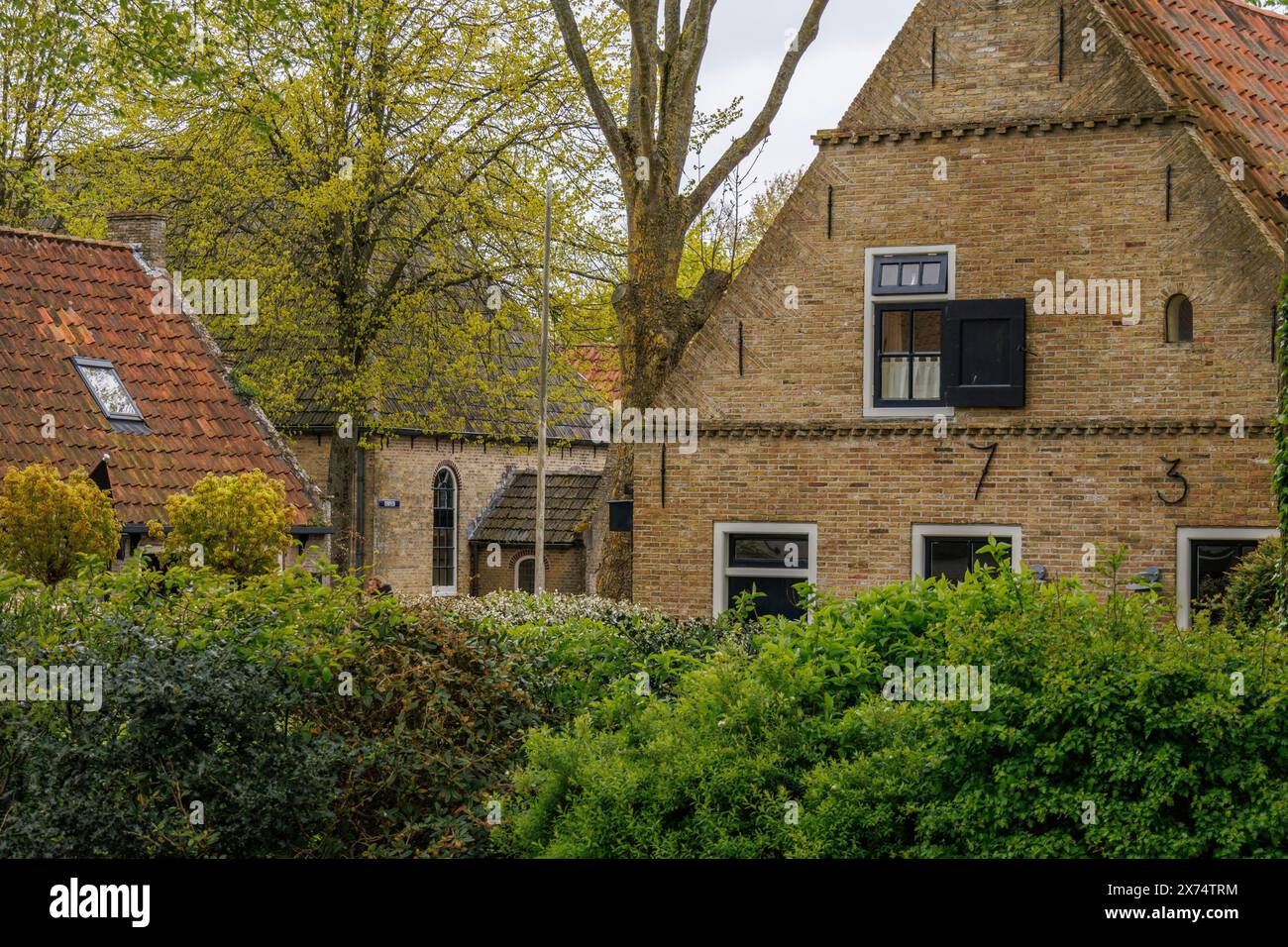 Ein altes Backsteinhaus mit großen Fenstern und umliegenden Bäumen und Sträuchern, alte Häuser mit grünen Gärten in einem kleinen Dorf, nes, ameland, The Stockfoto