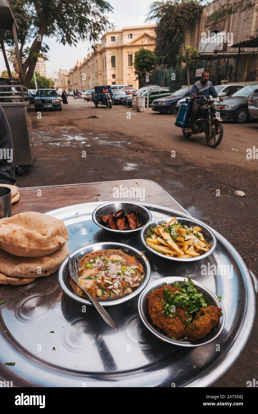 Ägyptisches Frühstück am Straßenrand im Zentrum von Kairo mit Falafel, Medames, Pommes, Auberginen und Baladi-Brot, serviert auf einem Metalltablett in der Nähe geparkter Autos Stockfoto