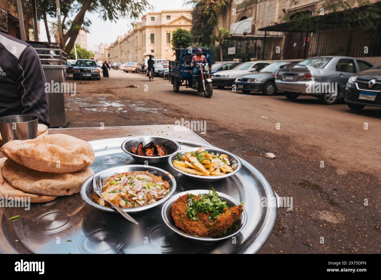 Ägyptisches Frühstück am Straßenrand im Zentrum von Kairo mit Falafel, Medames, Pommes, Auberginen und Baladi-Brot, serviert auf einem Metalltablett in der Nähe geparkter Autos Stockfoto