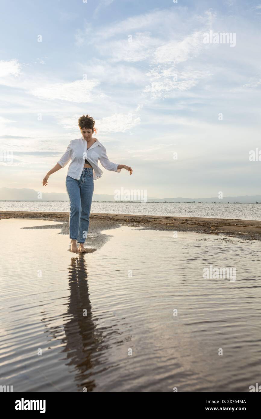 Frau am Strand tanzt bei Sonnenuntergang im Hintergrund mit der Sonne im Schatten, Frau im Urlaub, die auf dem Sand mit goldenen Wasserreflektionen f Stockfoto