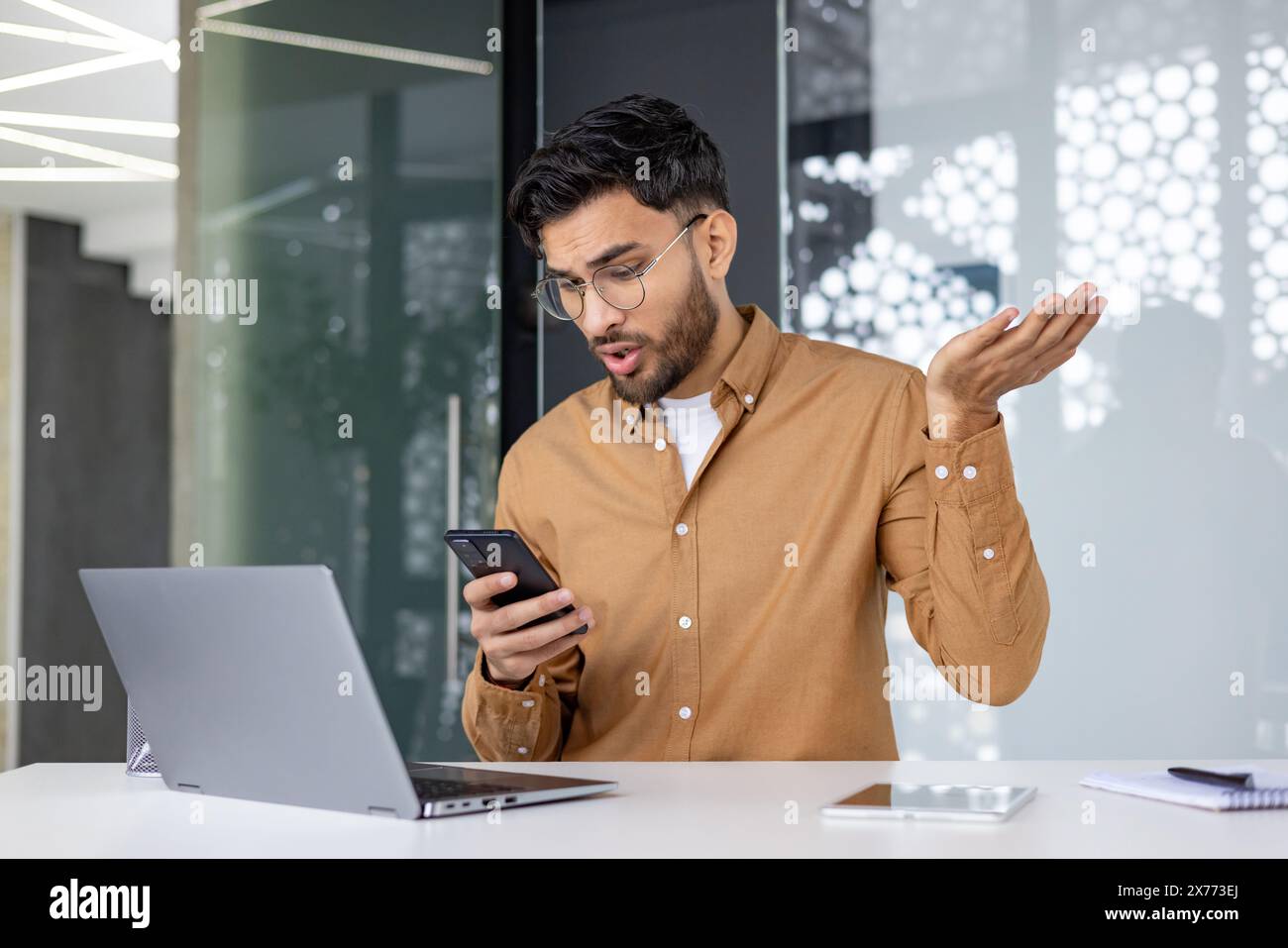 Büroangestellter sieht frustriert aus, als er schlechte Nachrichten auf seinem Handy liest, während er an seinem Laptop arbeitet. Stockfoto