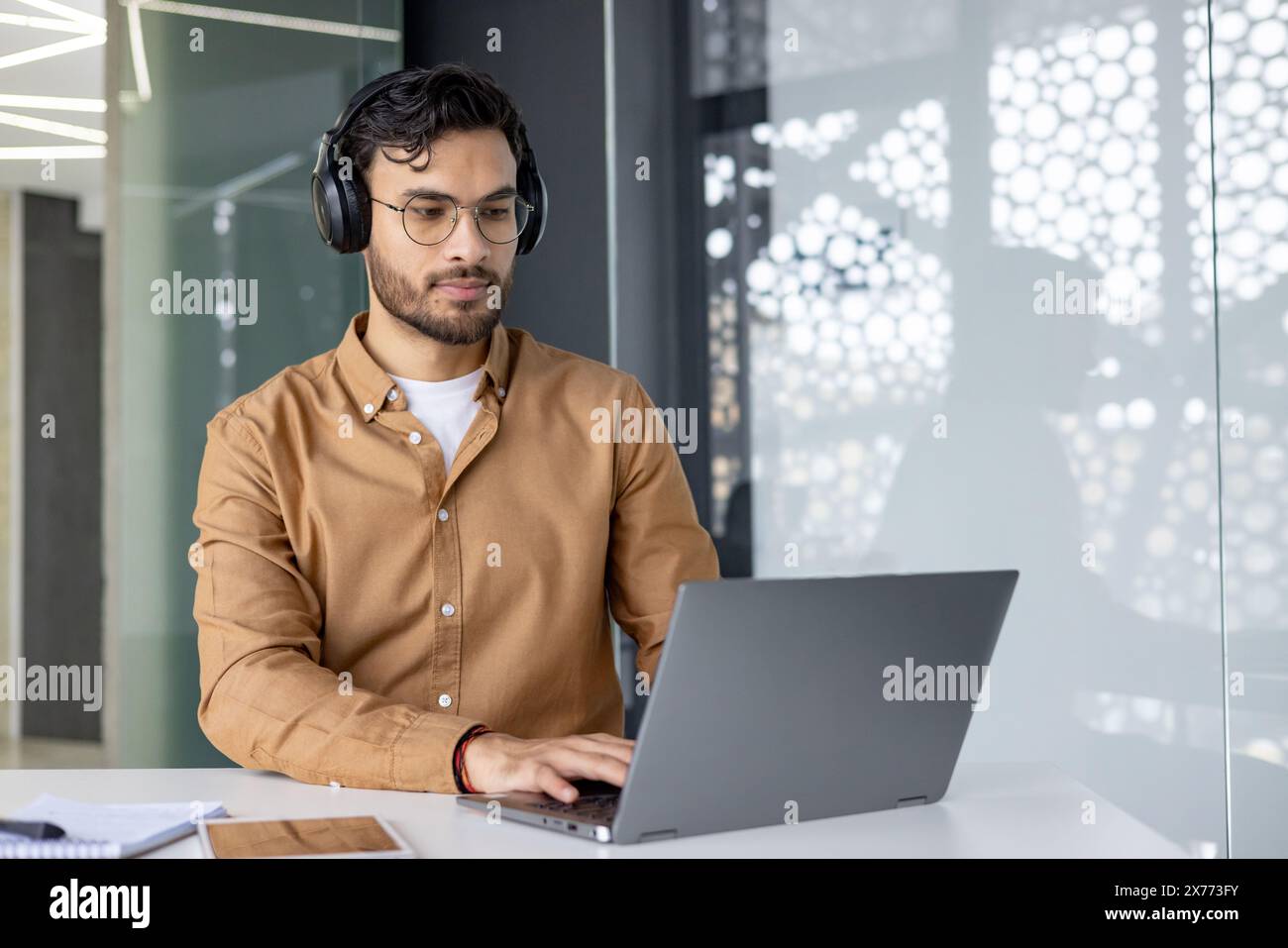 Ein fokussierter Mann mit Kopfhörern und Brillen, der an einem Laptop in einer modernen Büroumgebung arbeitet. Er konzentriert sich auf seine Aufgabe mit einem Notizblock und Tablet auf dem Tisch. Stockfoto