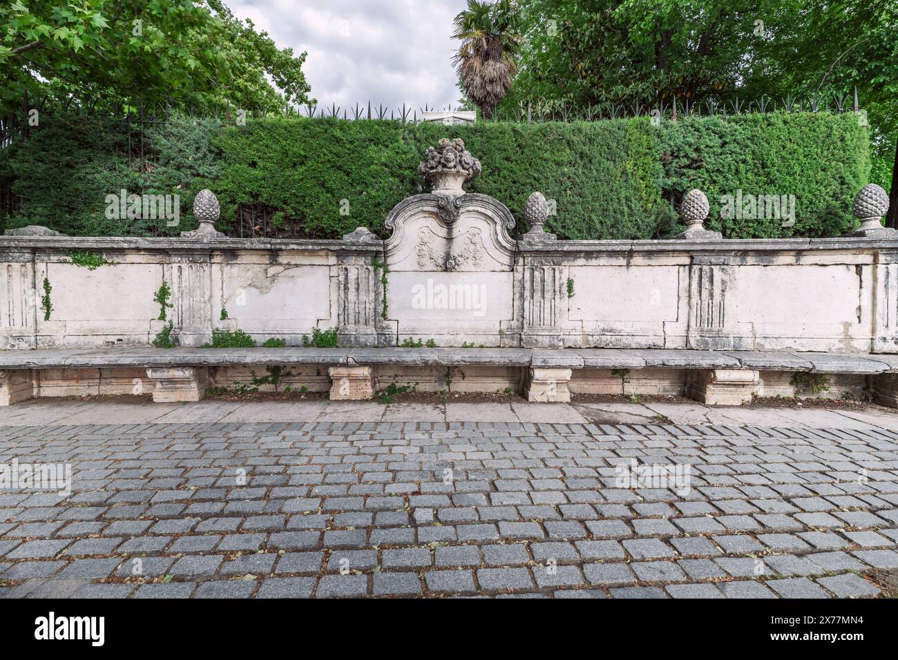 Eine kunstvolle antike Steinbank in einem Park mit einer Hecke auf der Oberseite und Steinpflasterböden Stockfoto