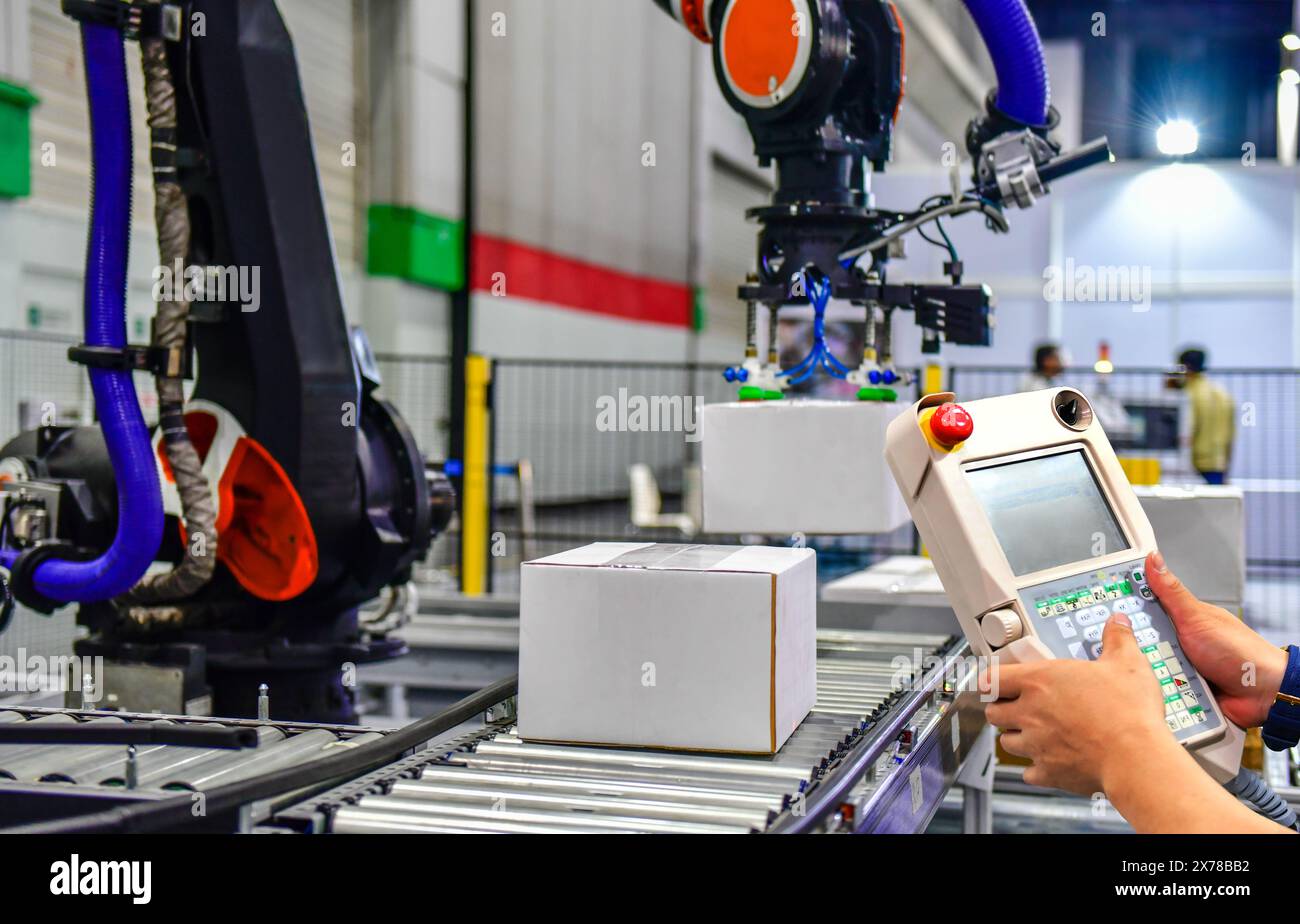 Der Manager prüft und steuert die Roboterarme der Automatisierung, die ein weißes Feld halten, und der Mitarbeiter, der eine Robotermaschine mit einem Bedienfeld bedient Stockfoto