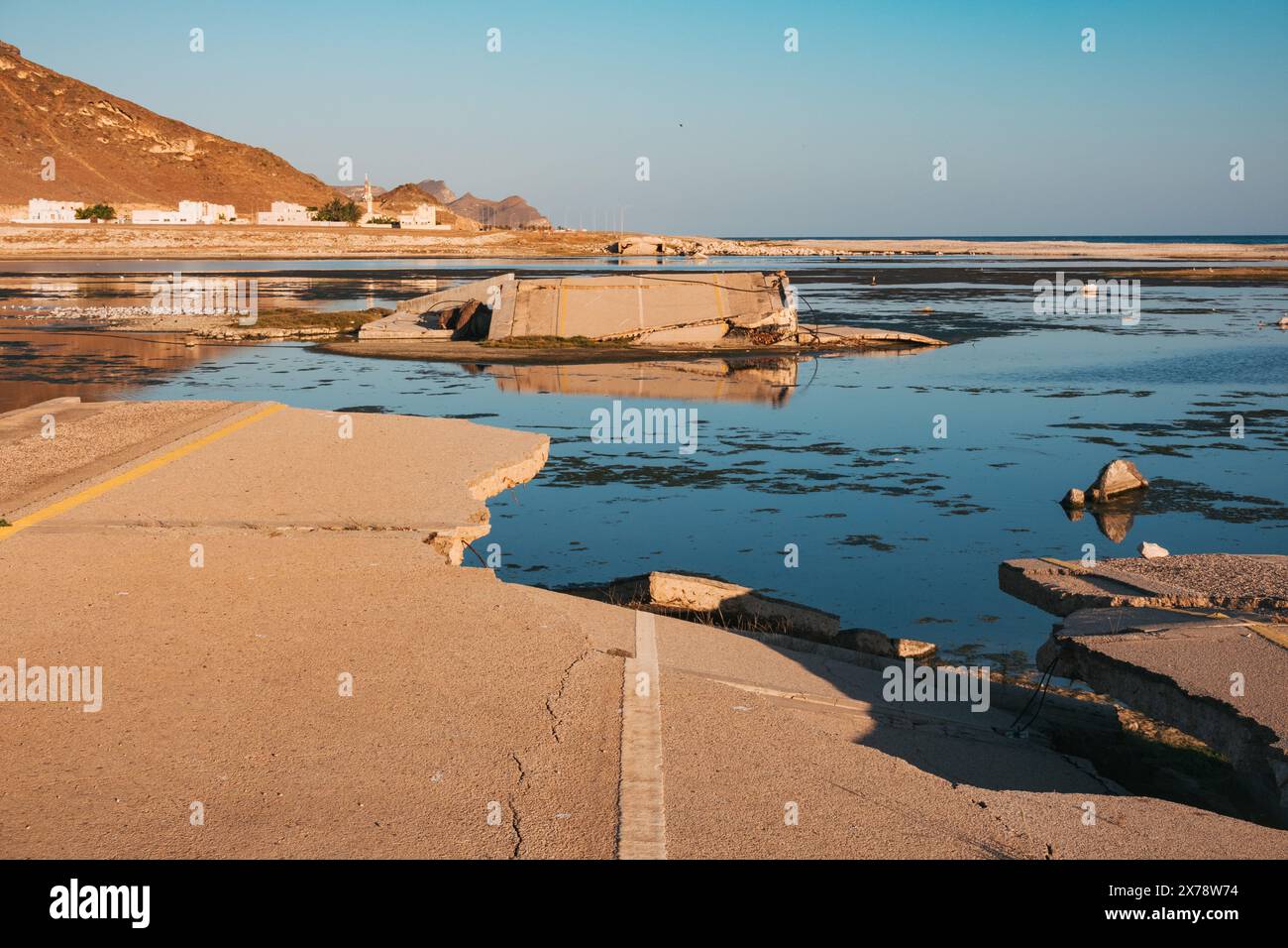 Eingestürzte Brücke am Al Mughsail Beach in Oman, mit abgebrochenen Straßenabschnitten, die vor einer Bergkulisse in seichtes Wasser führen. Stockfoto