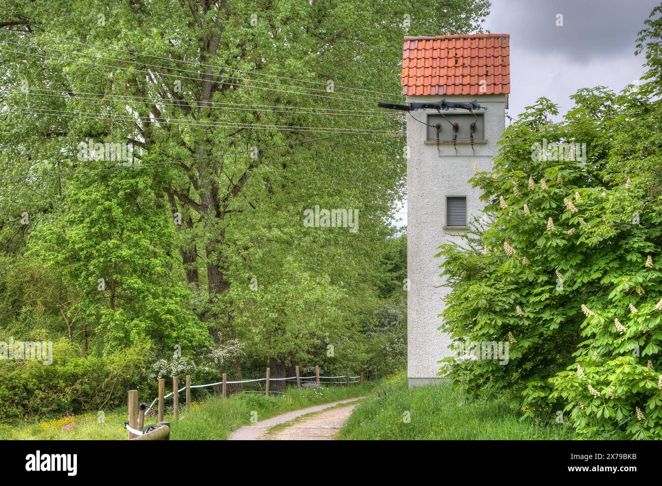 Ein einsamer Transformatorturm aus Stein steht inmitten von üppigem Grün, umgeben von einer malerischen Landschaft mit Wiesen und Bäumen entlang eines kleinen Kiesweges. Stockfoto