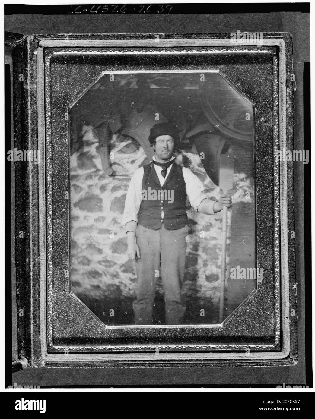 Berufsbildnis eines nicht identifizierten Mannes, in voller Länge, nach vorne gerichtet, mit einem langstieligen Werkzeug, vor einer Maschine und einer Steinmauer, Fotograf unidentifiziert., Hallmark: Rinhart 29., Fall: Vögel und Weinrebe., Kauf/Austausch; Gary Vroegindewey; 1981; (DLC/PP-1981:380R.6)., ist Teil von: Daguerreotype-Sammlungsarbeitern, 1850-1860. Stockfoto