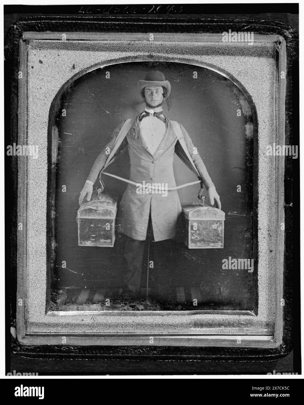 Berufsbildnis eines Händlers, durchgehend stehend, nach vorne gerichtet, mit zwei Taschen, die von einem Gurtzeug an den Seiten gehalten werden, Nackenstütze zwischen den Beinen sichtbar, Beschriftung aus der Ausstellung „American Treasures Memory“: Tools of the Trade“. Louis-Jacques-Mandé Daguerre erfand das Daguerreotypverfahren, das später in Frankreich am 19. August 1839 angekündigt wurde. Obwohl es sich um einen komplexen und langsamen Prozess handelte, nutzten amerikanische Fotografen diese neue Erfindung schnell, weil sie in der Lage waren, ein „wahrhaftiges Bild“ zu erfassen. Typischerweise Porträtaufnahmen in der Natur, Berühmtheiten, politischen Persönlichkeiten sowie Handwerker wir Stockfoto