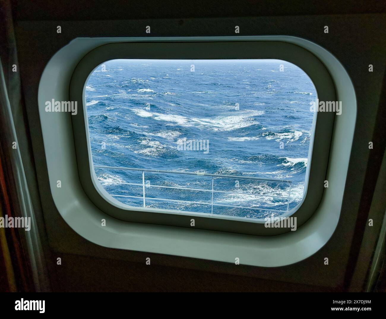 Bullaugenfenster Blick auf den Nordpazifik von Deck 8 aus gesehen. Die Wellen haben weiße Kappen, die raue See zeigen. Die Schiene ist unten zu sehen. Stockfoto