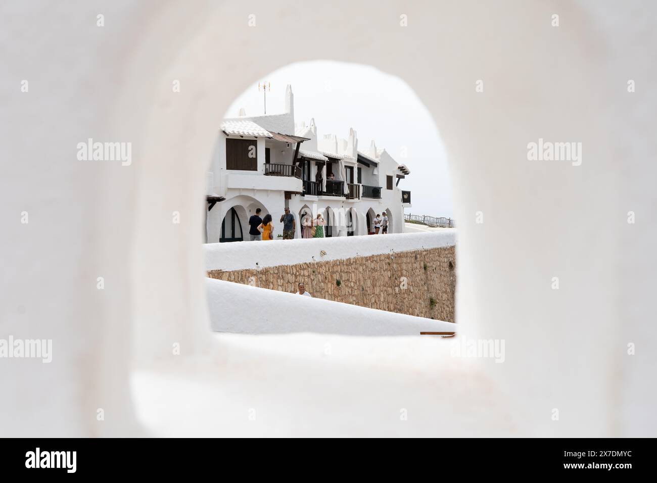 Eine typische Straßenszene in Binibeca, Menorca, mit Blick durch ein kleines Fenster eines weiß getünchten Hauses. Die bezaubernde mediterrane Architektur mit Menschen Stockfoto