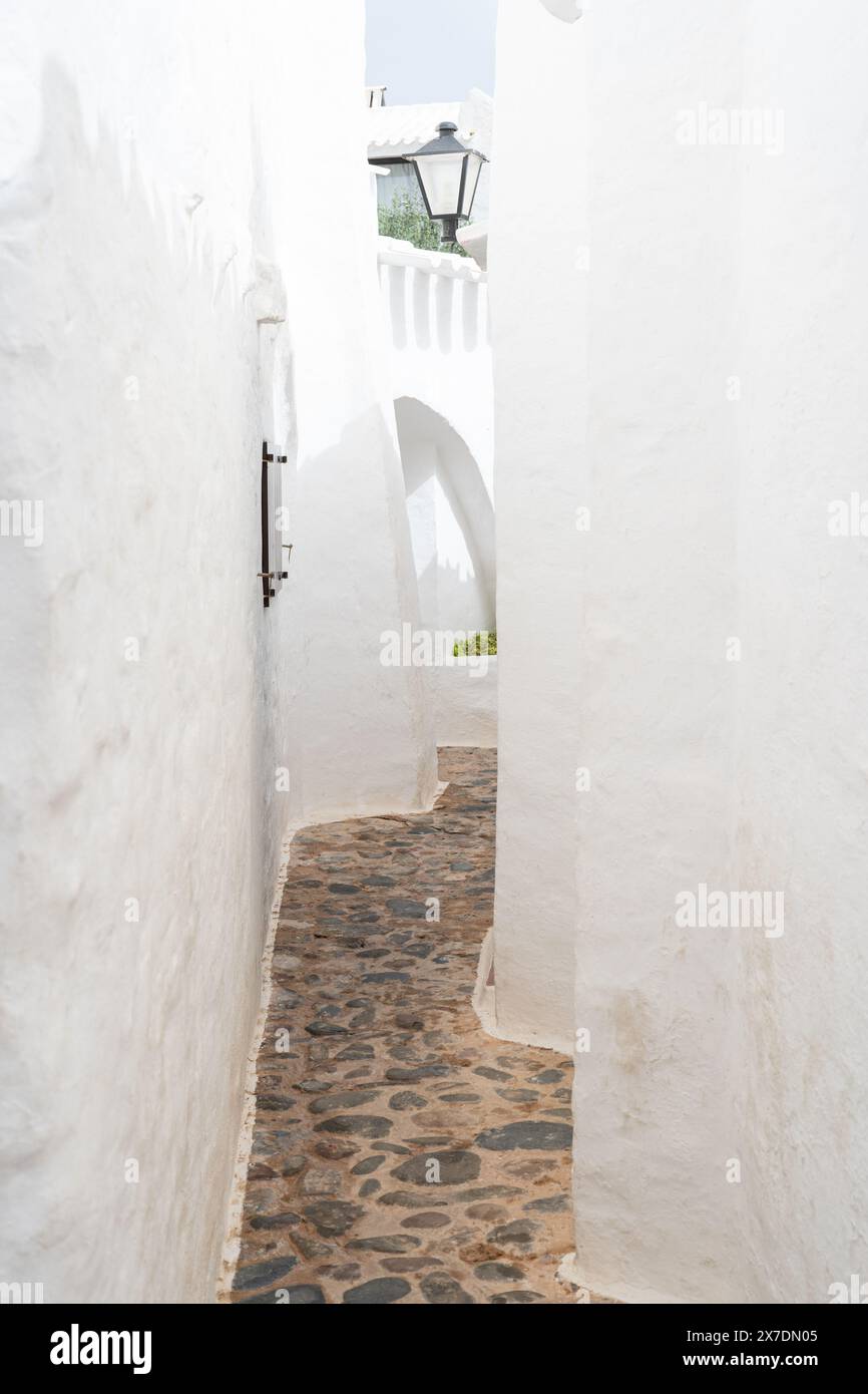 Ein malerischer Blick auf eine leere Straße in Binibeca, Menorca. Die weiß getünchten Gebäude und der kopfsteingepflasterte Pfad zeigen den bezaubernden mediterranen Bogen Stockfoto