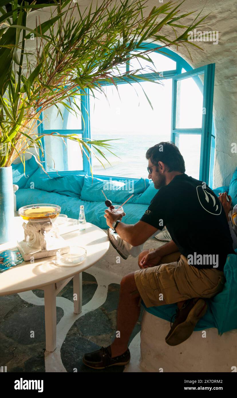 Mykonos, Griechenland - 11. Mai: Ein einsamer Mann an der Bar trinkt einen Drink, während er durch das große Fenster auf der Insel Mykonos die Aussicht draußen beobachtet Stockfoto