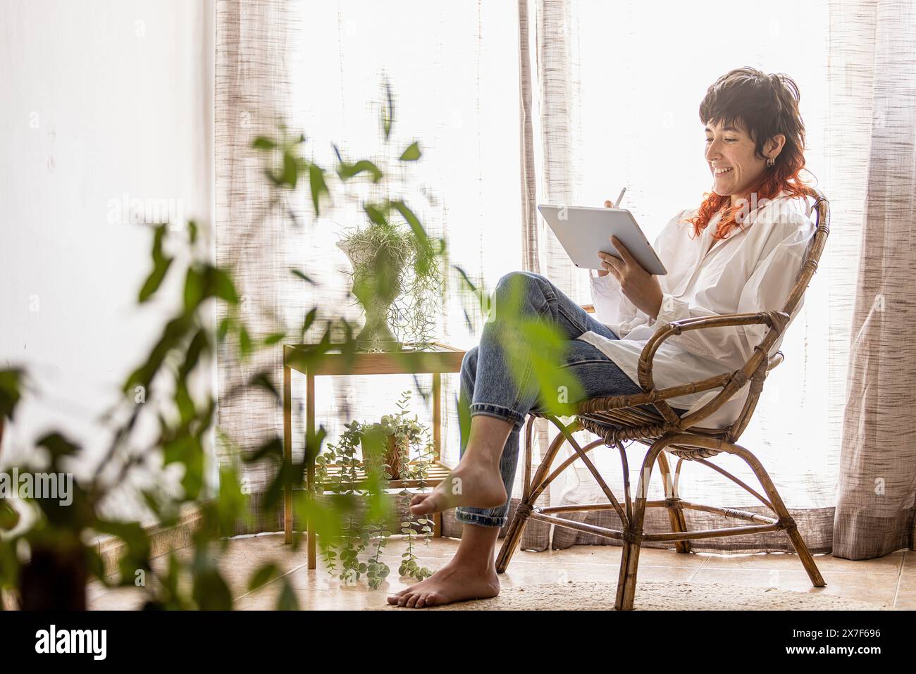 Frau, die zu Hause mit einem Tablet arbeitet schönes Fensterlicht gemütlich, lächelnd in die Kamera schaut, Kamera zeigt, Pflanzen Stockfoto