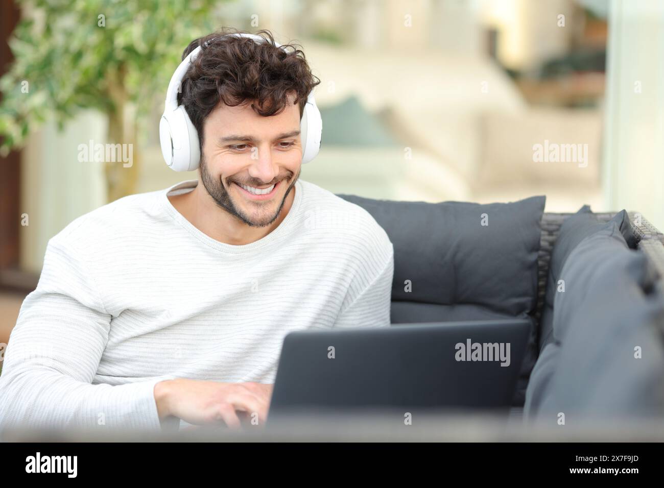 Glücklicher Mann mit Kopfhörern, der Medien auf dem Laptop ansieht und auf einer Couch auf einer Hausterrasse sitzt Stockfoto