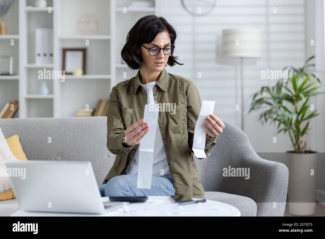 Besorgte und ernste junge Frau sitzt zu Hause auf dem Sofa, überprüft Belege für Einkäufe und Ausgaben, mit Laptop und Rechner. Stockfoto