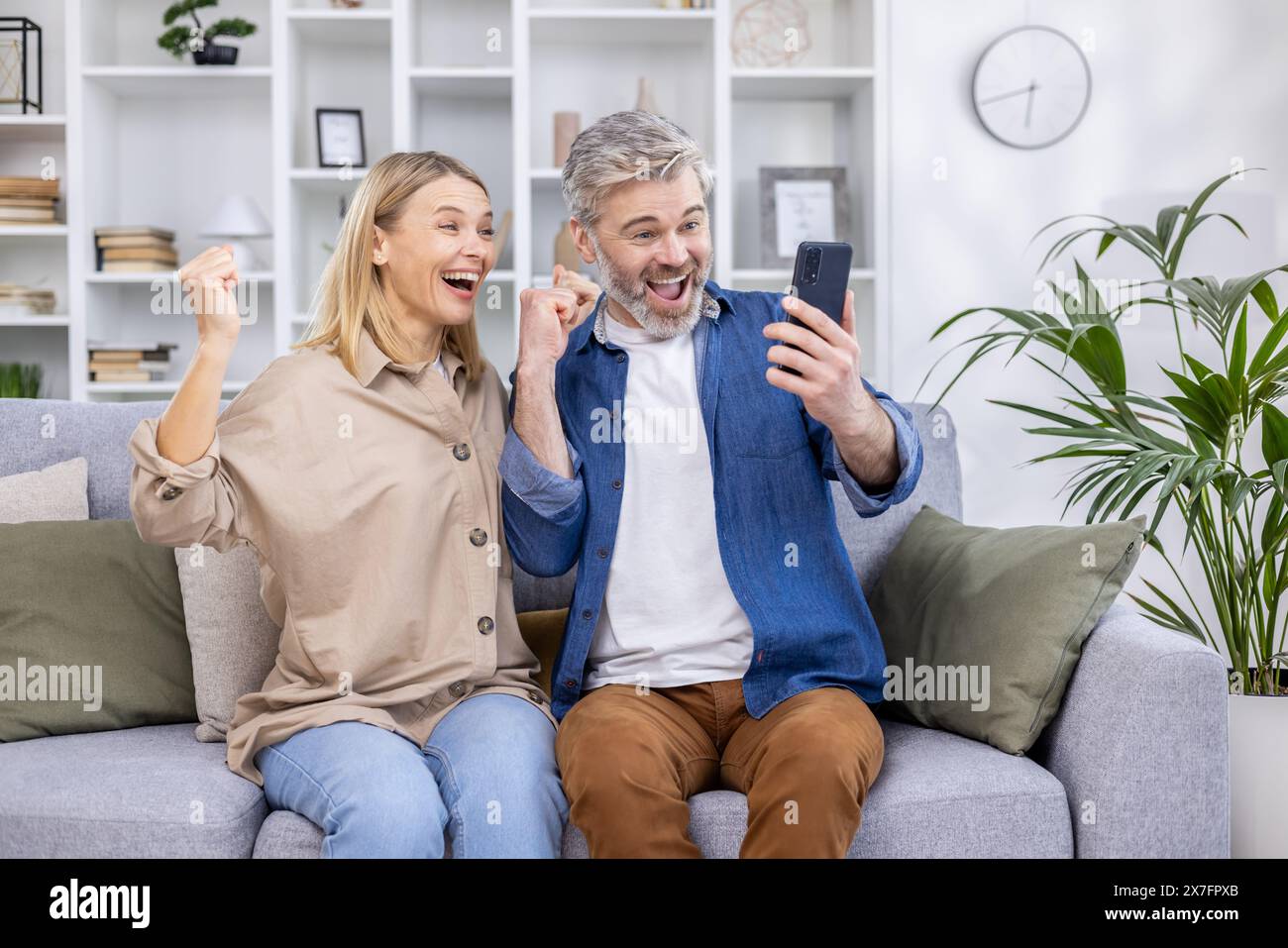 Älteres Paar feiert freudig Erfolg, während es auf das Smartphone blickt, Aufregung und Glück zeigt und auf einer Couch in einem hellen Wohnzimmer sitzt. Stockfoto