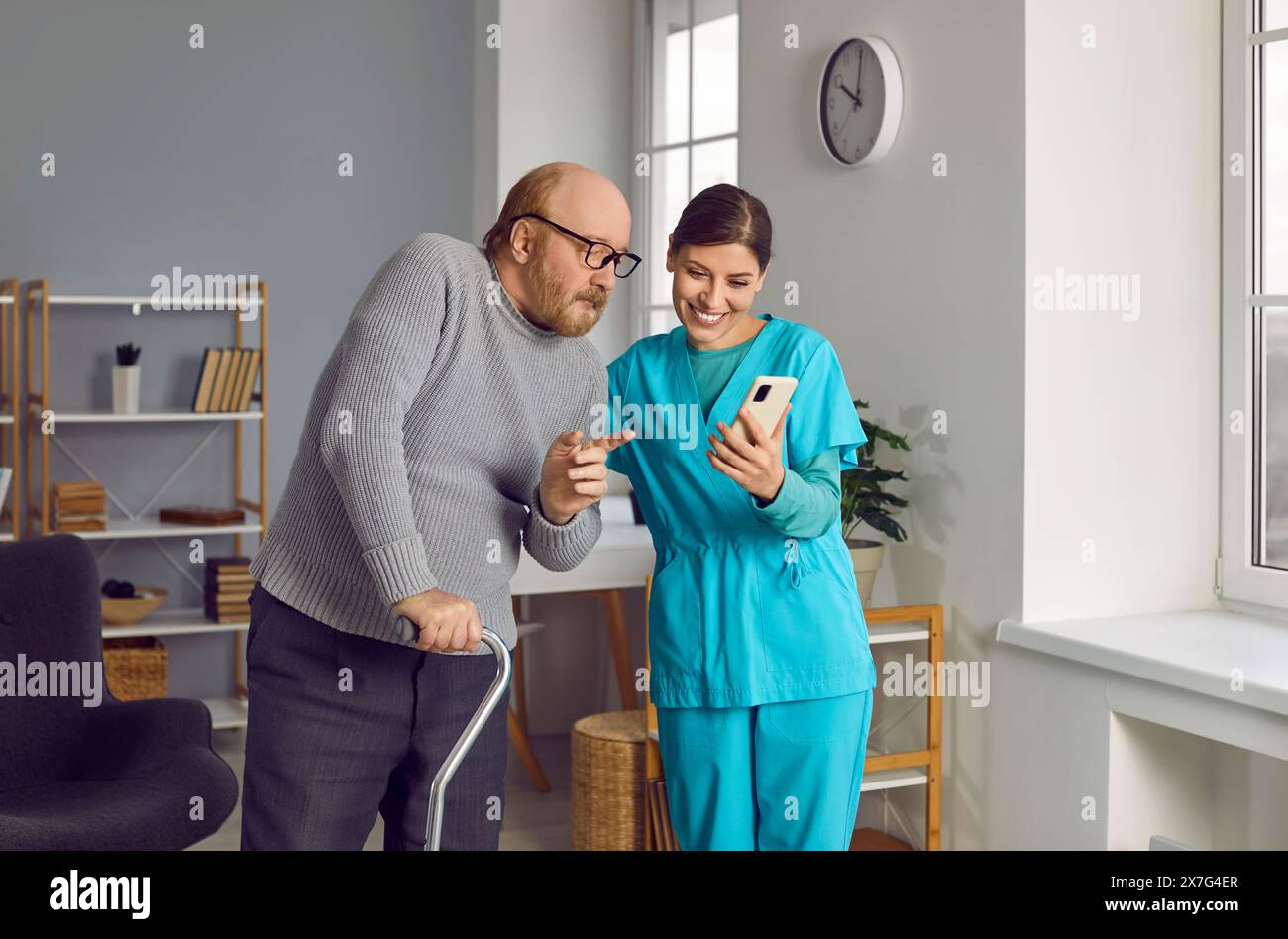 Bärtiger, rothaariger Rentner im warmen Pullover zeigt auf den Handy-Bildschirm seiner jungen, lächelnden Schwester. Pflege der älteren Generation in der Krankenpflege Stockfoto