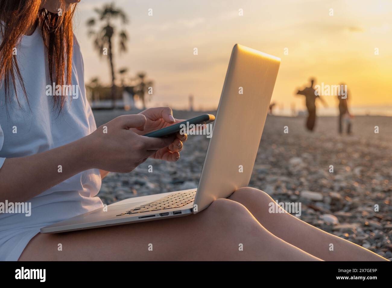 Nahaufnahme einer Frau, die mit Laptop und Handy am Strand am Meer während des Sonnenuntergangs arbeitet. Freiberufliche Frauen arbeiten online mit dem Smartphone und Stockfoto