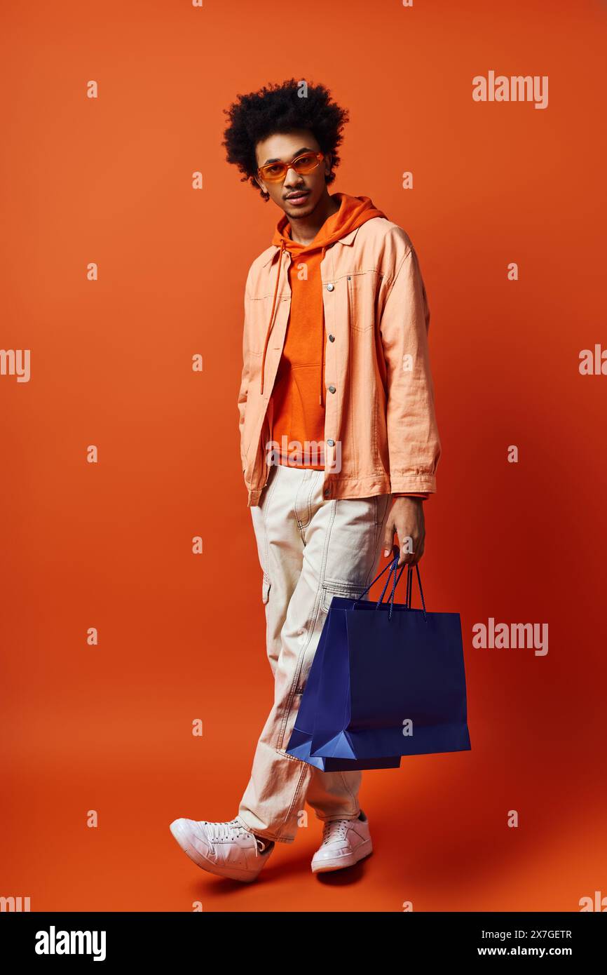 Ein junger Afroamerikaner in einem leuchtend orangefarbenen Hemd und einer weißen Hose, der eine blaue Tasche vor einem leuchtend orangefarbenen Hintergrund hält. Stockfoto