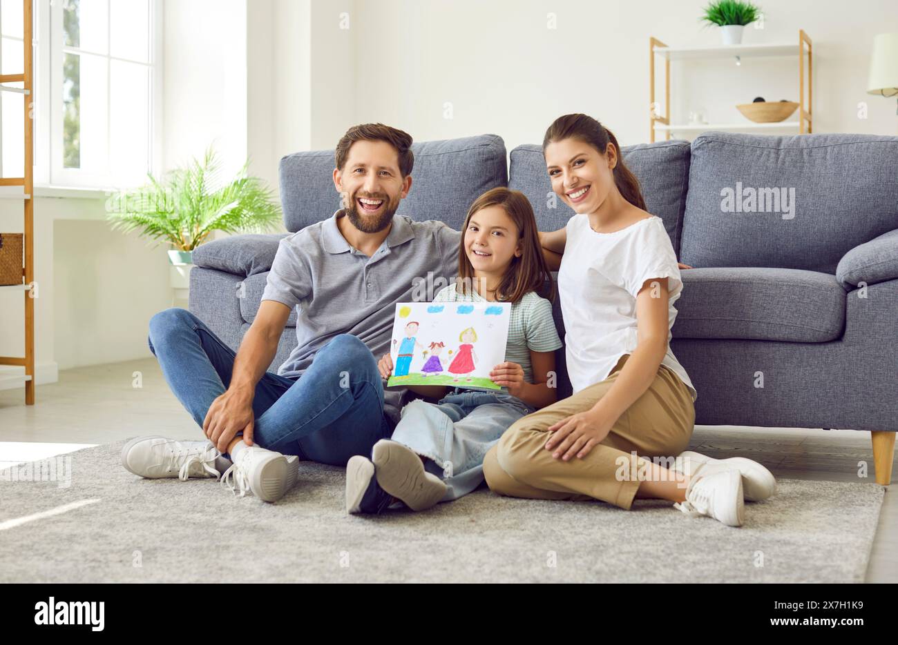 Lächelndes Mädchen, das auf dem Boden sitzt und die Eltern zu Hause zeigen, wie eine glückliche Familie gezeichnet ist. Stockfoto