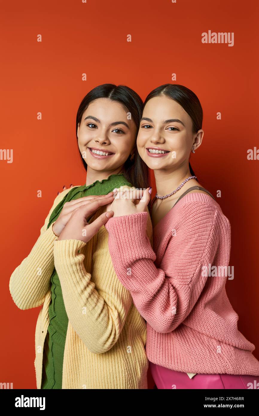 Zwei hübsche und brünette Teenager-Mädchen in legeren Kleidern stehen nebeneinander vor einer auffälligen roten Wand in einem Studio. Stockfoto