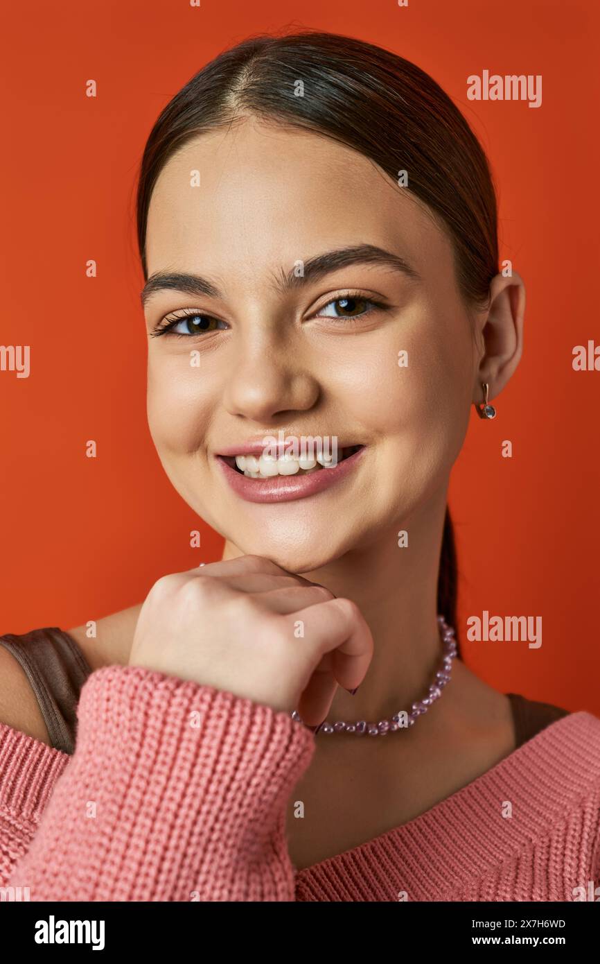 Ein brünettes Mädchen im Teenageralter lächelt in einem rosa Pullover und strahlt Freude und Glück in einem Studio mit orangem Hintergrund aus. Stockfoto