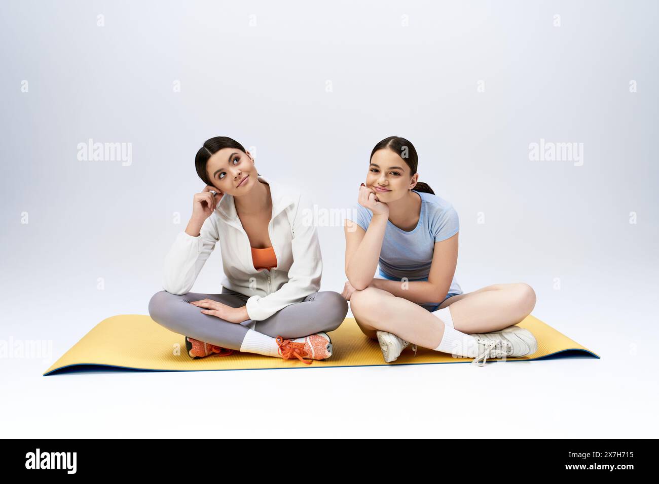 Zwei hübsche, brünette Teenager-Mädchen in sportlicher Kleidung sitzen auf einer Yogamatte und teilen einen Moment der Entspannung und Verbindung. Stockfoto