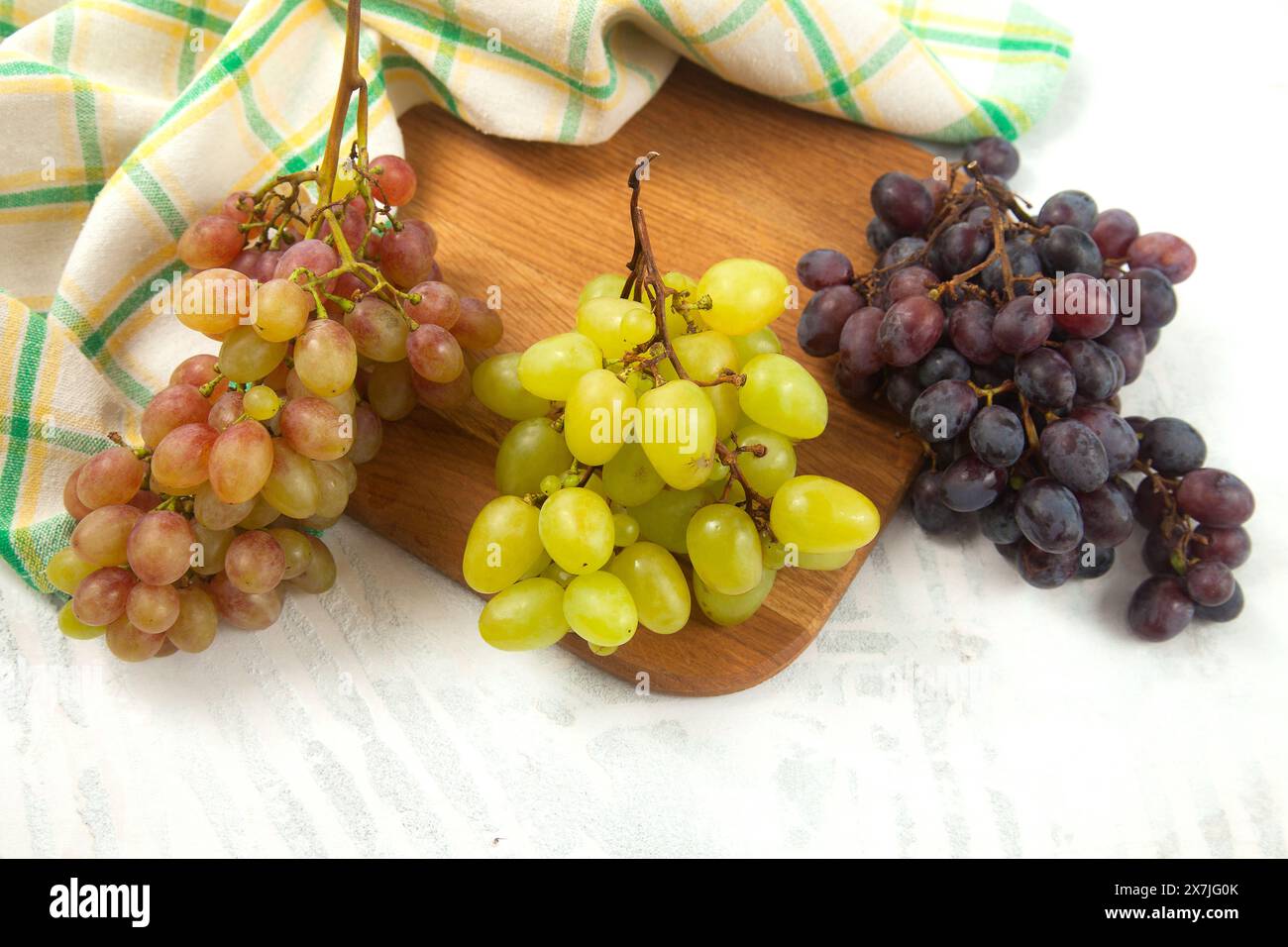 Schneidebrett mit einem großen Haufen reifer grüner, rosa und dunkelblauer Trauben auf weiß. Weintrauben mit grünem Küchentuch auf weißem hölzernem Hintergrund. Stockfoto