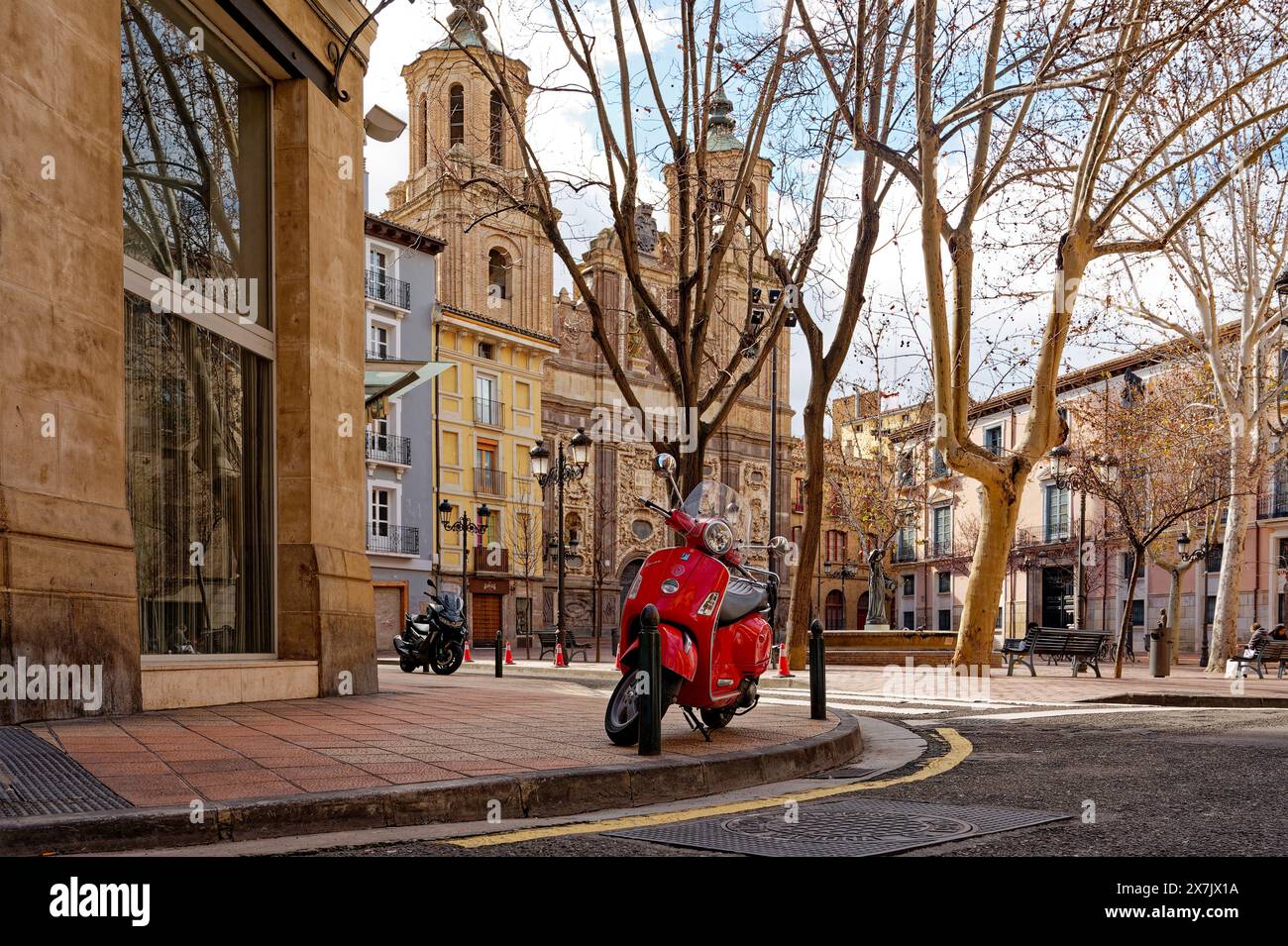 Rotes klassisches Motorroller-Motorrad, das an einer Ecke neben einem baumlosen platz mit der barocken Kirche Santa Isabel de Portugal geparkt ist. Stockfoto