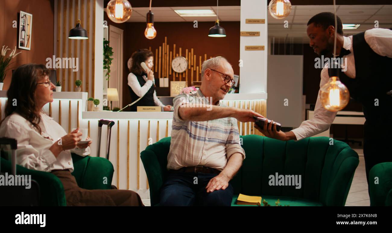 Pensioniertes Paar zahlte für eine Kaffeetasse von der Hotelbar und nahm Kreditkarte für POS-Terminal-Transaktion. Ehemann und Ehefrau entspannen sich gemeinsam im Loungebereich des Resorts und zahlen für den Service. Stockfoto