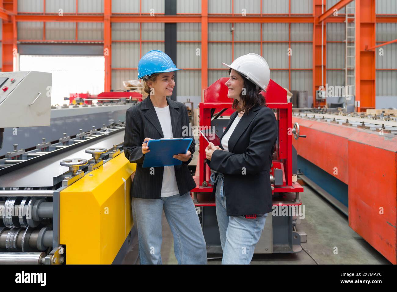 Zwei Frauen mit Schutzhelm und schwarzem Anzug stehen in einer Fabrik, halten ein Klemmbrett und einen Tablet-Computer, benutzt um Notizen zu machen, den Zeitplan zu überprüfen, r Stockfoto