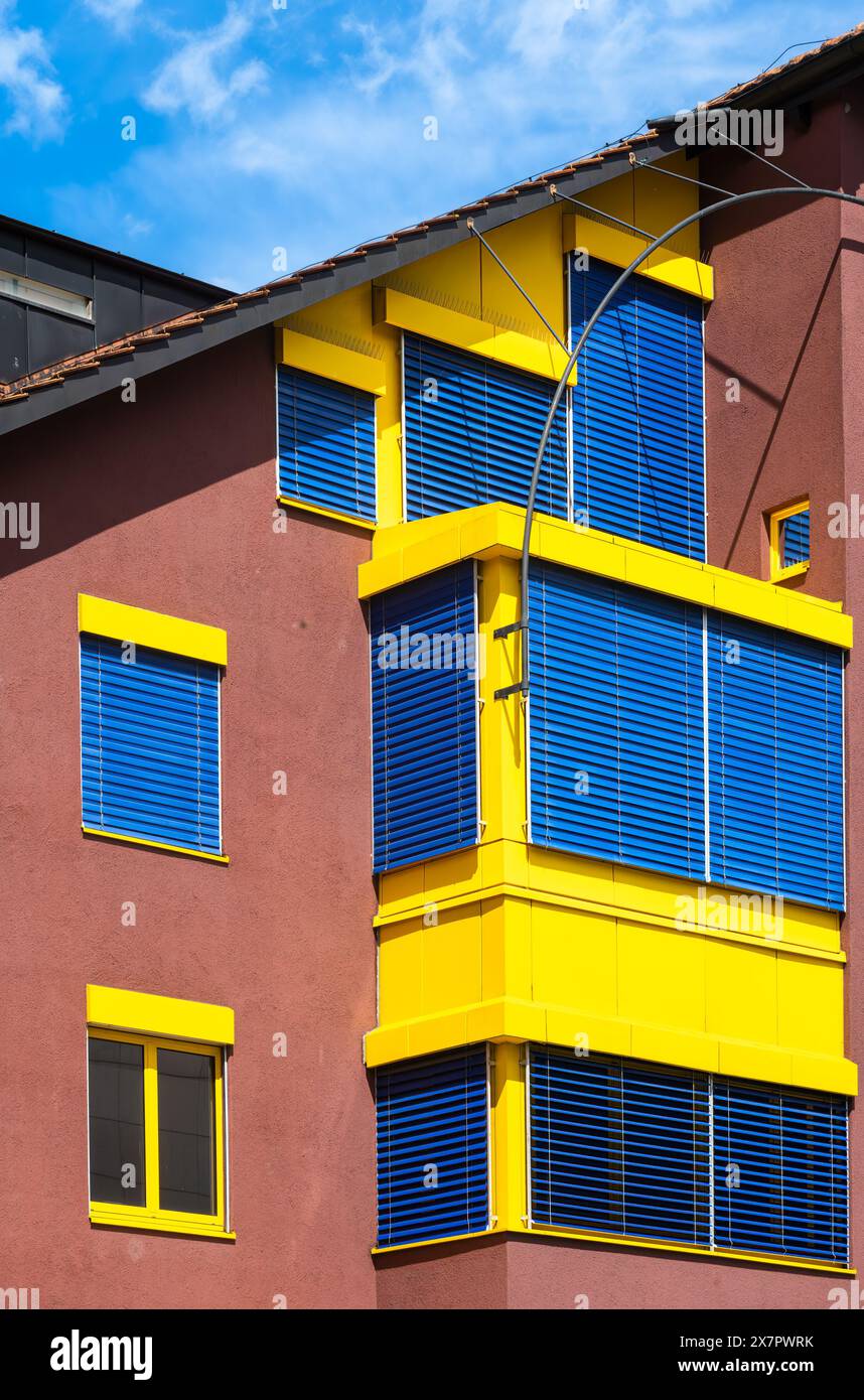Schaan, Liechtenstein - 17. Mai 2024: Ein lebendiges, modernes Gebäude mit einer satten braunen Fassade, gelben Fensterrahmen und blauen Jalousien unter klarem Himmel, Stockfoto