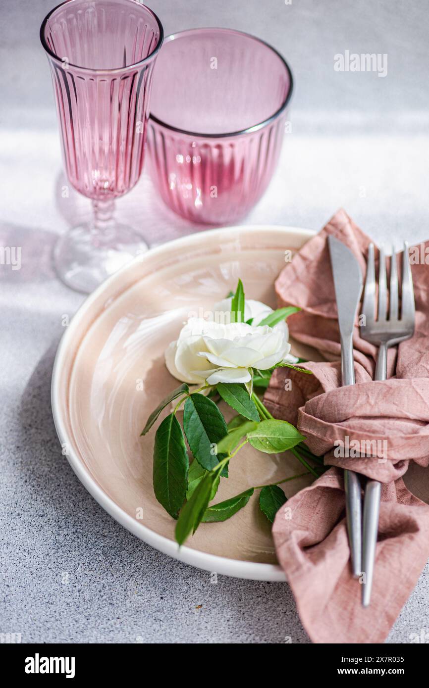 Ein eleganter Tisch mit einem rosafarbenen Pastellteller, silbernem Besteck in eine dunkel rosa Serviette und weißer Rose neben rosafarbenem g Stockfoto