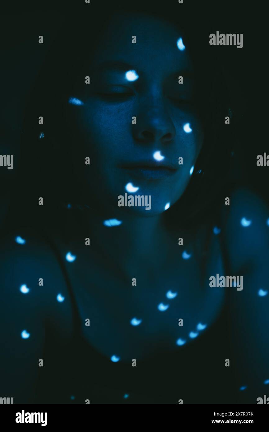 Eine ruhige Frau, die von kleinen leuchtenden Lichtpunkten bedeckt ist, wird in einem dunklen Raum gefangen, der von blauem Licht beleuchtet wird und eine traumhafte, reflektierende Stimmung verkörpert Stockfoto