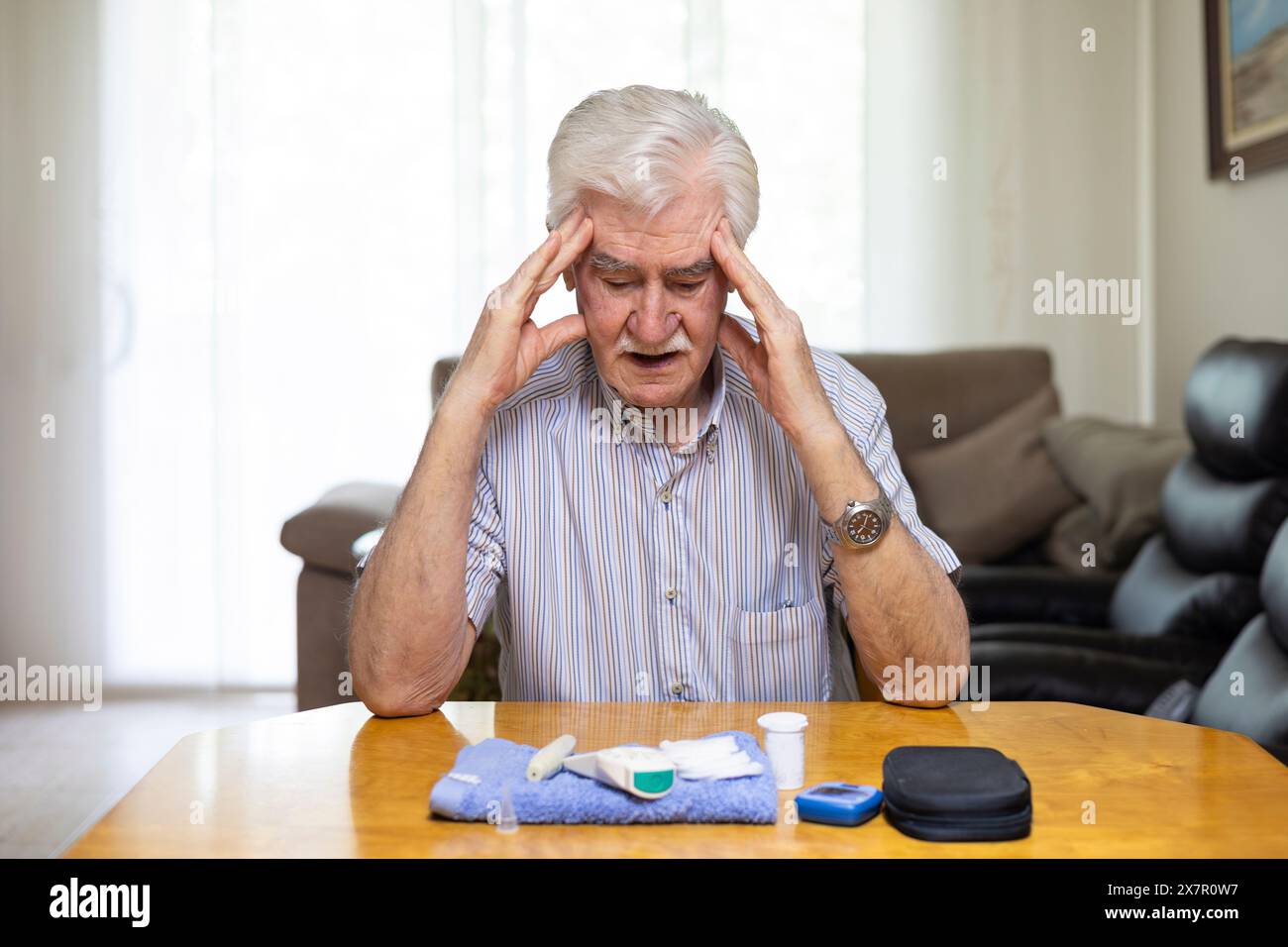 Ein älterer Mann sitzt an einem Tisch und fühlt sich aufgrund eines niedrigen Blutzuckerspiegels unwohl medizinische Geräte und Medikamente sind im Vordergrund sichtbar, Highlightin Stockfoto