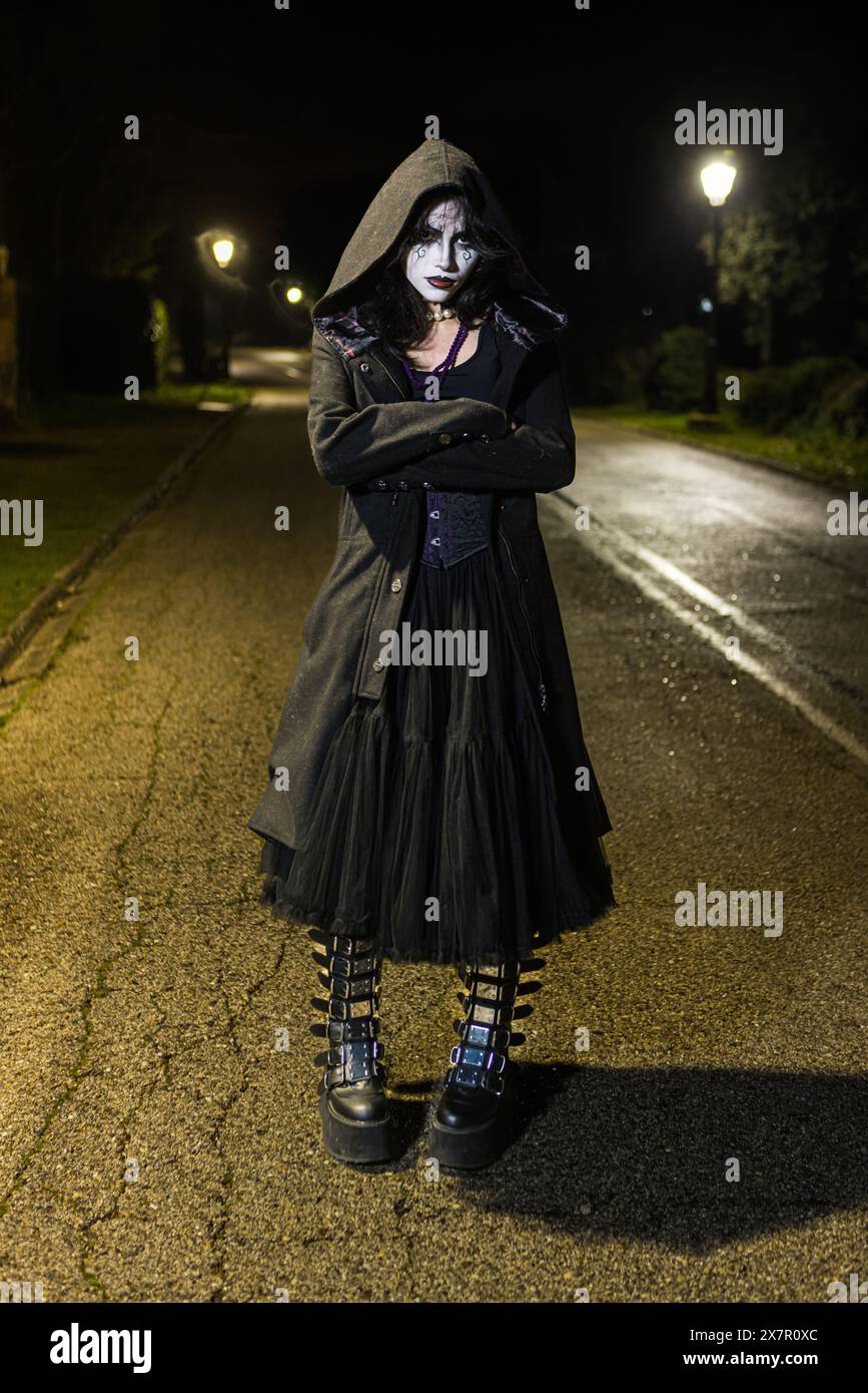 Eine Frau mit dramatischem Gothic-Make-up steht nachts auf einer schlecht beleuchteten Straße und blickt direkt in die Kamera, mit einer dezenten Feststimmung, die an erinnert Stockfoto