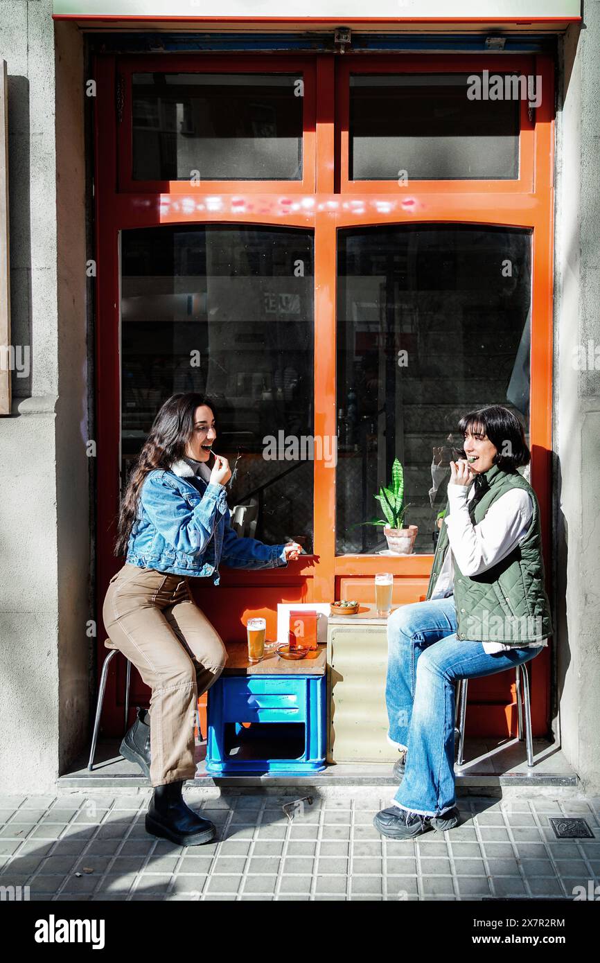 Weibliches Paar, das einen sonnigen Tag zusammen in einem Café im Freien genießt und sich mit fröhlichen Ausdrücken unterhalten hat. Stockfoto