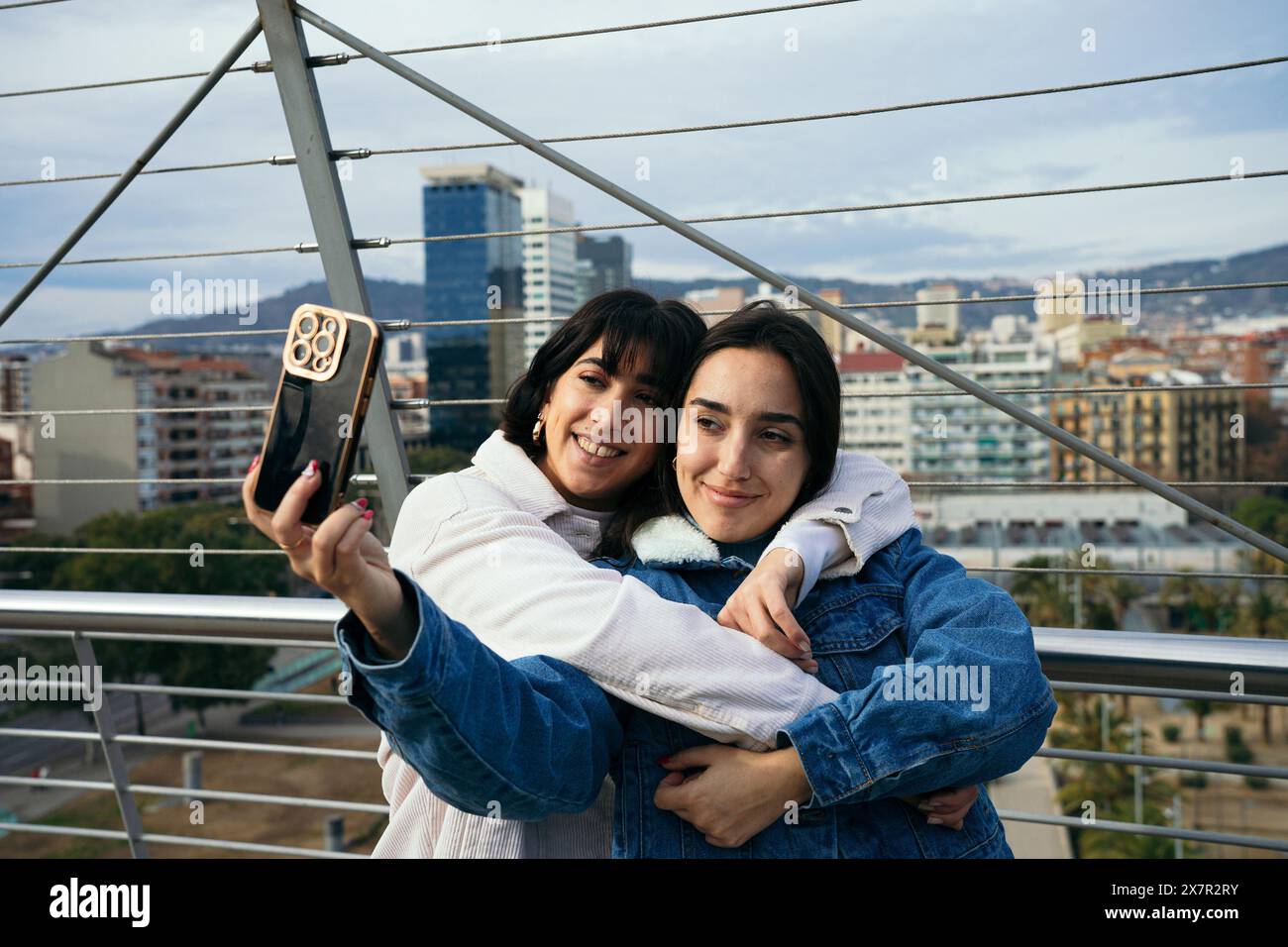 Weibliches, fröhliches Paar umarmt sich während eines Selfie mit einem Smartphone auf einem Dach der Stadt und teilt einen glücklichen Moment. Stockfoto