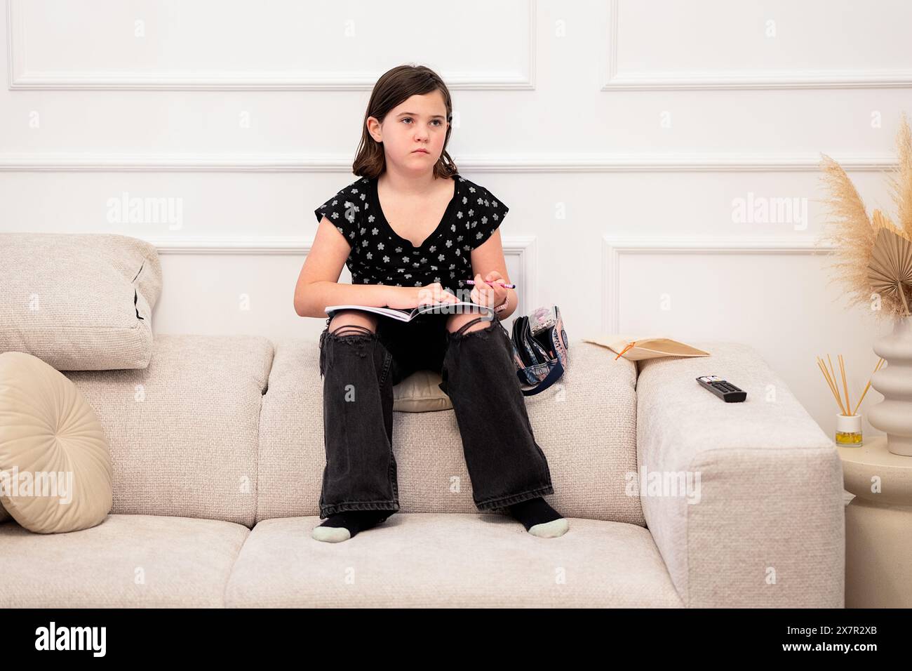 Ein junges Mädchen sitzt zu Hause bequem auf einem beigen Sofa, vertieft in ein Buch mit nachdenklichem Look, trägt legere Kleidung und blickt weg von der CA Stockfoto
