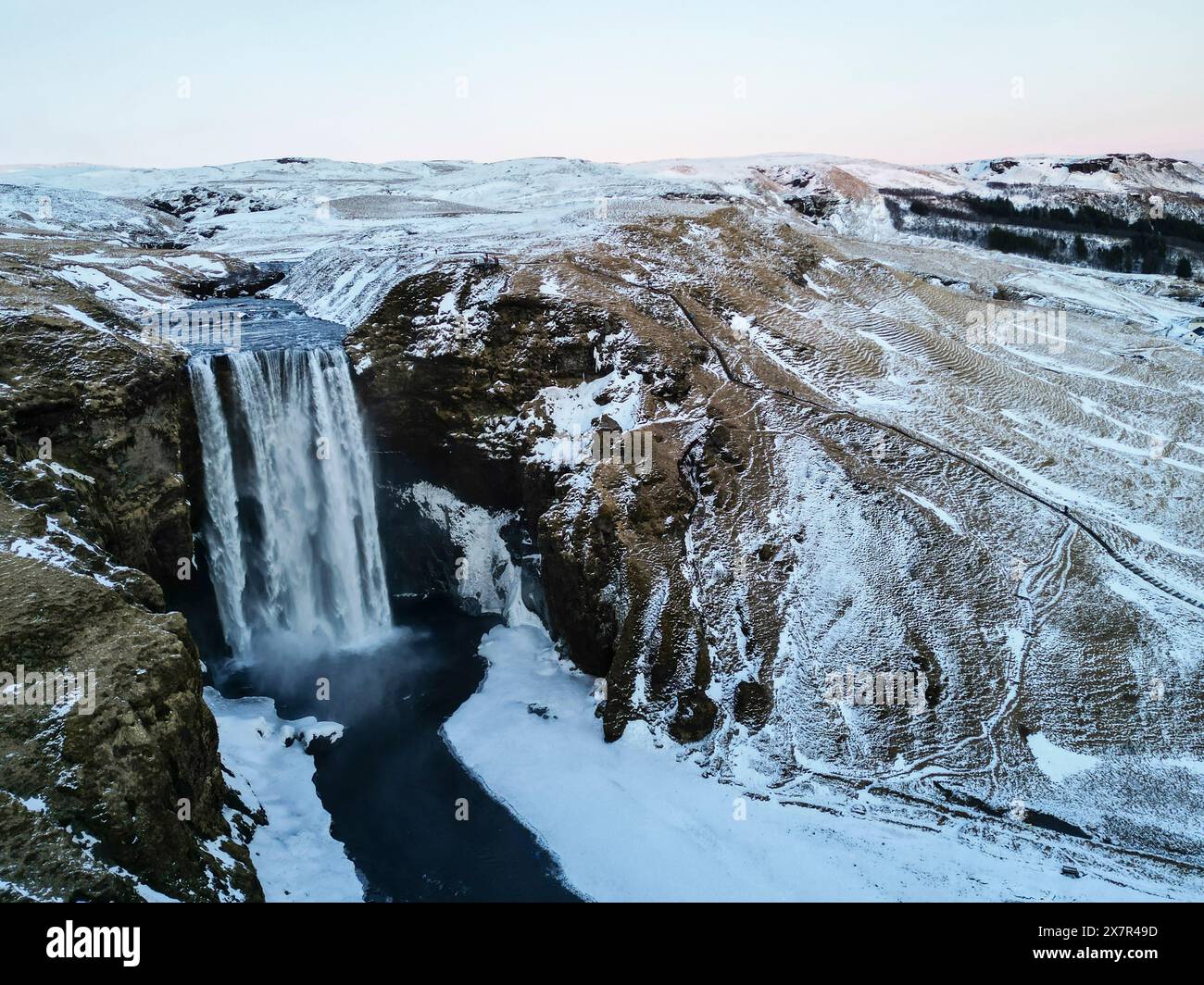 Eine atemberaubende Luftaufnahme eines mächtigen Wasserfalls, der inmitten schneebedeckter Klippen im zerklüfteten Gelände Islands sprudelt Stockfoto