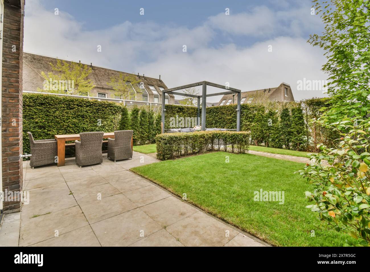 Ein üppiger Vorstadtgarten mit einem geräumigen Rasen, Der von dichten Hecken umgeben ist. Ein Essbereich mit Tisch und Stühlen ist teilweise mit modernem Metall bedeckt Stockfoto