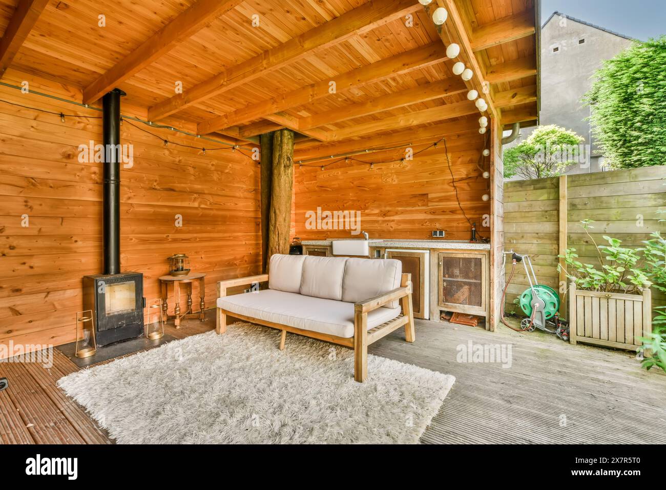 Eine warme und einladende Holzterrasse mit modernem Sofa, Teppich, Lichterketten und einem Holzofen für Komfort. Stockfoto