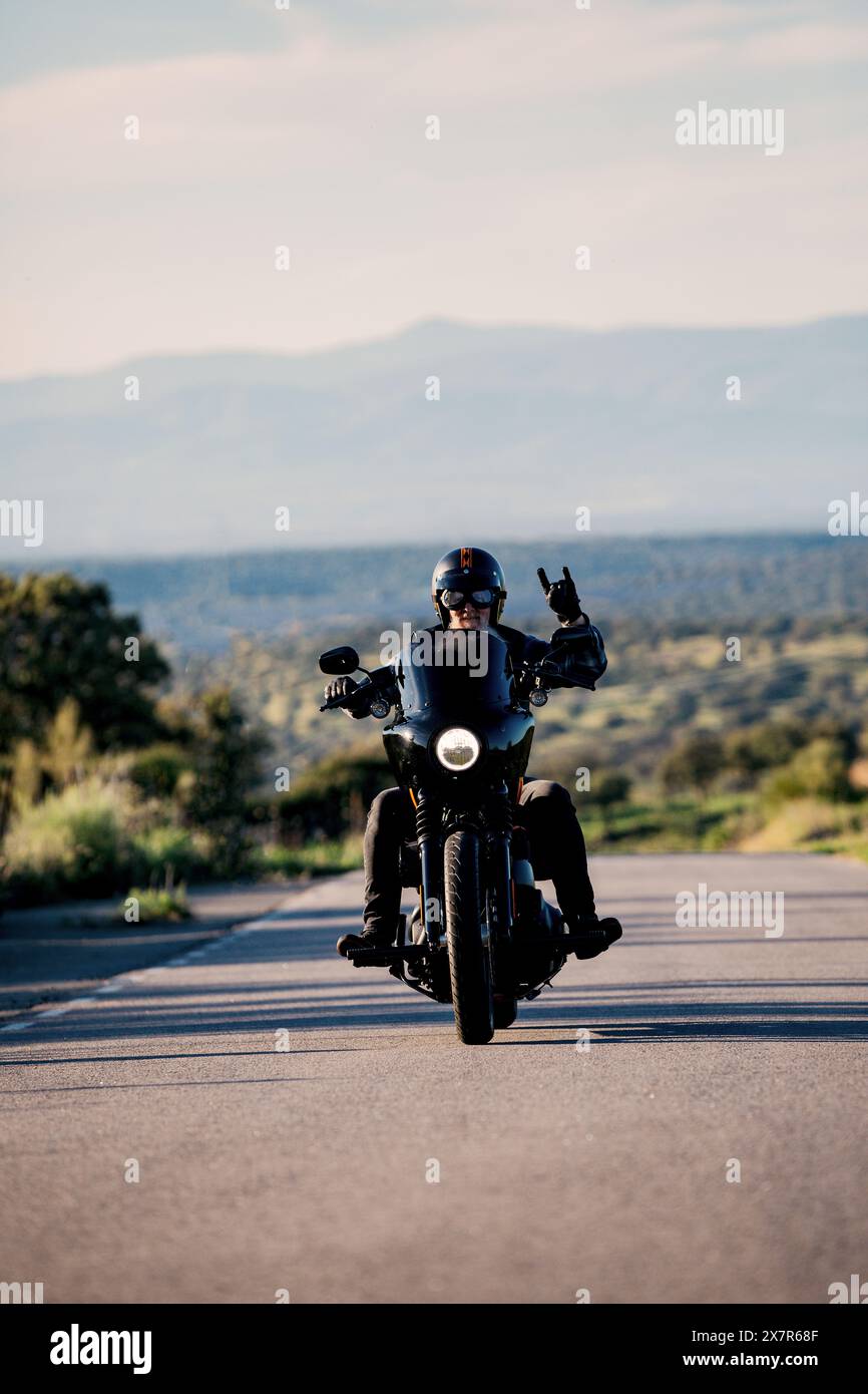 Ein Motorradfahrer in Schutzkleidung fährt auf einer offenen Straße in einer ruhigen Landschaft, macht ein Friedenszeichen, blickt nach vorne, nicht in die Kamera. Stockfoto