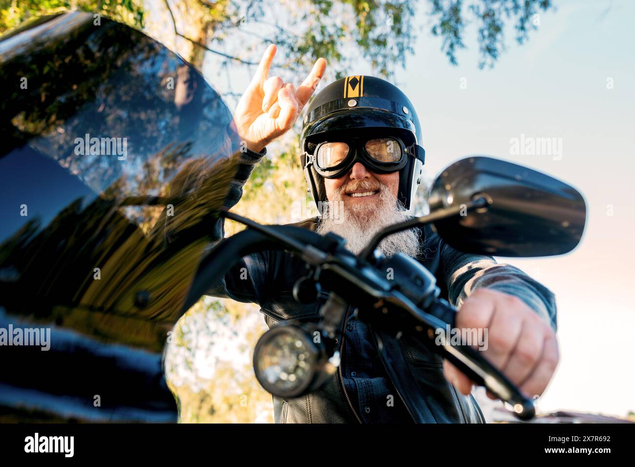 Von unten gibt ein fröhlicher Seniorenmann mit Bart und Helm ein Friedenszeichen, während er auf dem Motorrad fährt und in die Kamera blickt. Stockfoto