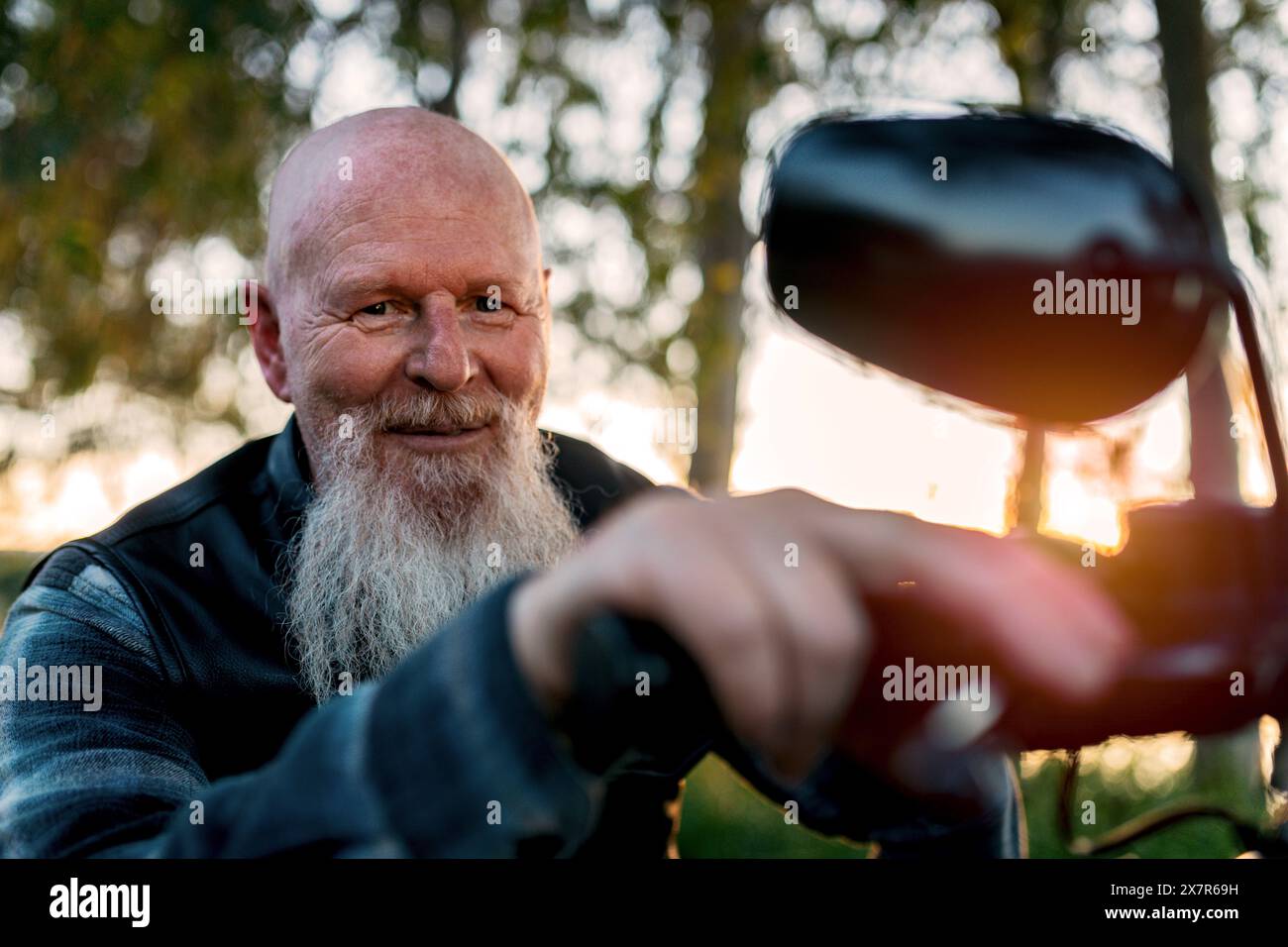 Ein fröhlicher älterer Mann mit weißem Bart und lächelndem Ausdruck sieht man, wie er den Seitenspiegel eines Motorrads bei Sonnenuntergang verstellt. Er schaut auf Cam Stockfoto