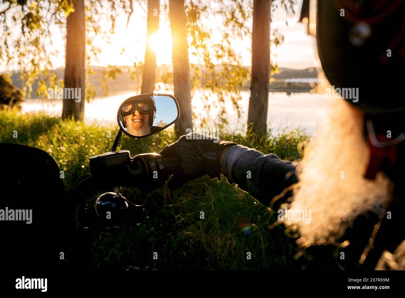 Ein lächelnder, bärtiger Seniorenmann mit Helm spiegelt sich im Seitenspiegel eines Motorrads wider und blickt auf die Kamera mit einem Hintergrund von sonnendurchfluteten Bäumen Stockfoto