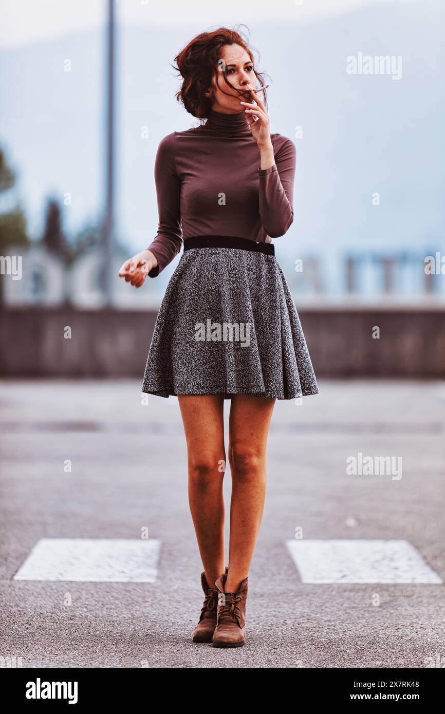 Frau, gekleidet mit braunem Rollkragen und grauem Rock, geht auf einer leeren Straße. Ihre entspannte Haltung und der ferne Blick deuten auf tiefe Gedanken hin. Der Hintergrund Stockfoto