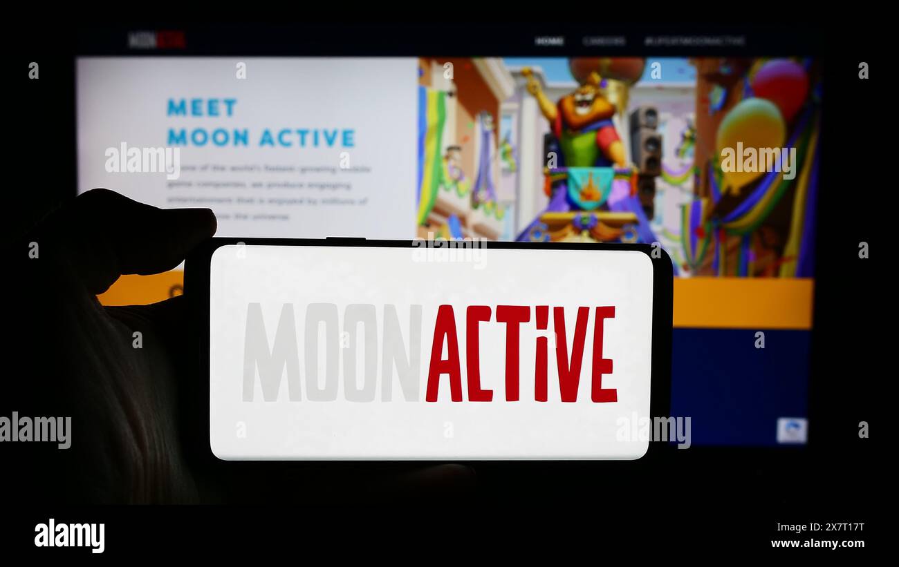 Person, die ein Mobiltelefon mit dem Logo des israelischen Spieleunternehmens Moon Active Ltd. Vor der Webseite hält. Schwerpunkt auf dem Display des Telefons. Stockfoto