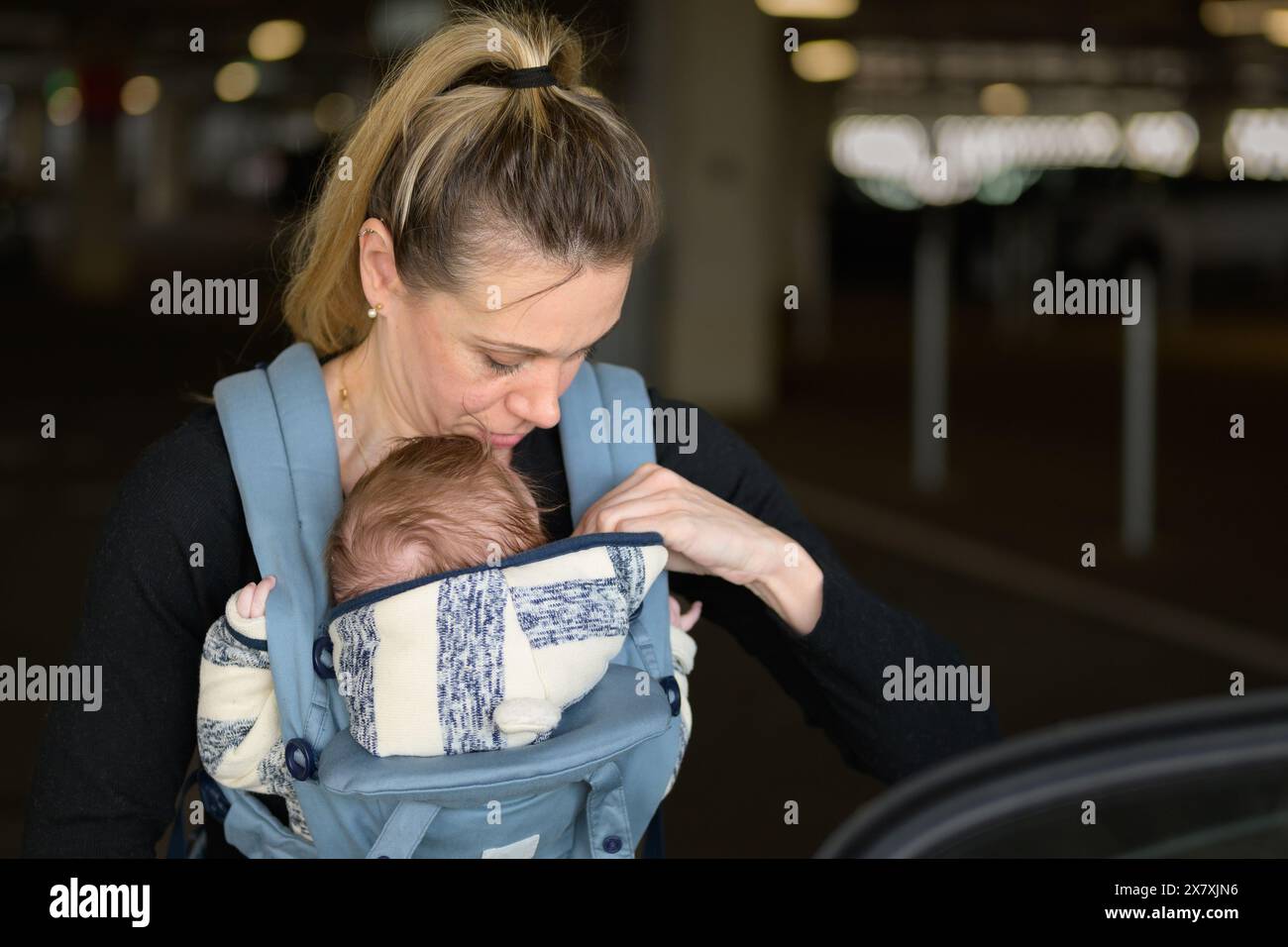 Porträt einer blonden Frau mittleren Alters, die prüft, ob ihr Baby gut sitzt, während es es in einer Babytrage auf einem Parkplatz hält und trägt Stockfoto